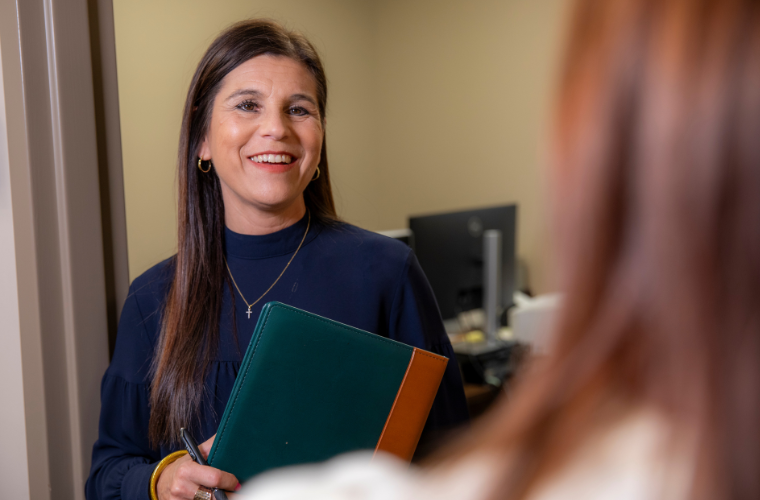 A woman is pictured standing in an office doorway holding a portfolio and speaking with someone off-camera.